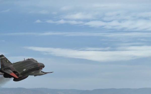 A military jet aircraft, appearing to be an F-4 Phantom or similar vintage fighter, is captured in low-level flight over what appears to be a desert military installation. The aircraft is in a banked turn with visible engine exhaust smoke trailing behind it. The background features arid mountainous terrain and ground-level military or industrial infrastructure.