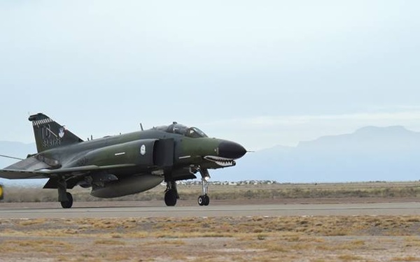 A military jet aircraft, identifiable as an F-4 Phantom II with a distinctive shark mouth nose art and olive drab camouflage paint scheme, is landing on a desert airstrip with its drag parachute fully deployed. The aircraft appears to be rolling out on the runway, using the chute for deceleration. The setting appears to be a arid, desert environment with mountains visible in the background.