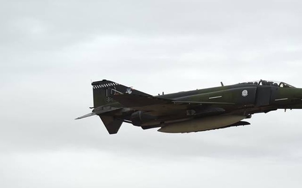 A military jet fighter aircraft is captured in level flight against an overcast sky. The aircraft features distinctive nose art including a shark mouth design and appears to be in a dark olive/green camouflage paint scheme. A checkered pattern is visible on the tail fin, and the aircraft carries external stores/ordnance under its fuselage.