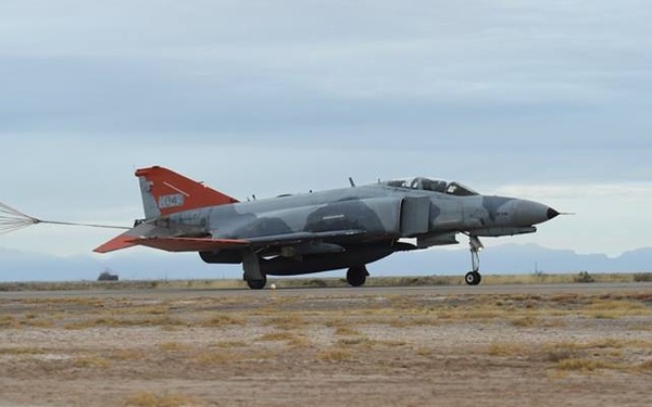A military jet aircraft, identifiable as an F-4 Phantom II, is landing on a desert airstrip with its drag parachute deployed to assist in deceleration. The aircraft features a gray camouflage paint scheme with distinctive red tail markings. The setting appears to be an arid, desert environment with mountains visible in the background.