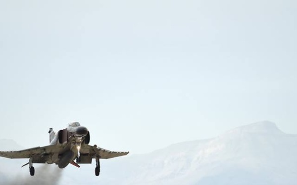 A military jet aircraft, consistent with an F-4 Phantom II, is captured at the moment of takeoff from a desert airstrip. The aircraft is in full afterburner, producing visible exhaust smoke and heat distortion behind it. The setting appears to be an arid, high-desert environment with mountains visible in the background.