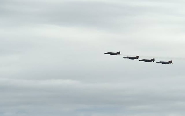 Four military jet aircraft are flying in a diagonal echelon formation against an overcast, grey sky. The aircraft appear as dark silhouettes at distance, flying from left to right over a hilly or mountainous horizon. The image captures a formation flight, likely a military exercise or ceremonial flyover.