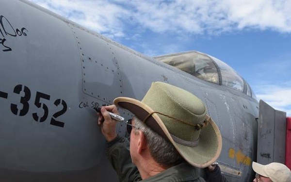 Two men are signing the fuselage of a military jet aircraft, identified as AF-352, on what appears to be an airfield tarmac. The aircraft bears existing signatures and markings, including 'PermDog' and '23TFS' written near the nose. One man wears a military-style flight jacket and wide-brimmed hat, while the other wears civilian attire with a badge/lanyard.