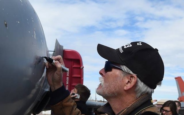 A middle-aged man wearing a black cap, sunglasses, and a brown jacket is writing or signing on the fuselage of a large gray military aircraft using a marker. He has a Nikon camera strap around his neck, suggesting he may be a photographer or civilian visitor. In the background, an orange-tailed F-16 fighter jet is visible on what appears to be a military airfield tarmac.