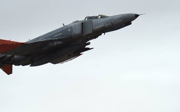 A military jet aircraft, identifiable as an F-4 Phantom II, is captured in flight against an overcast gray sky. The aircraft is painted in a dark gray/charcoal scheme with distinctive bright red markings on the tail section and wing tips, suggesting a drone or target aircraft configuration. The jet appears to be in a slight climbing or level flight attitude, photographed from a low angle.