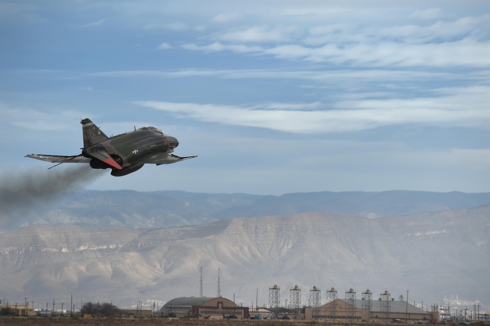 A military jet aircraft, identifiable as an F-4 Phantom II, is captured in a low-altitude, high-speed pass over what appears to be a military air base. The aircraft is in a banked turn with engine exhaust smoke visible, set against a desert landscape with mountains in the background. Ground infrastructure including hangars and antenna towers is visible below.