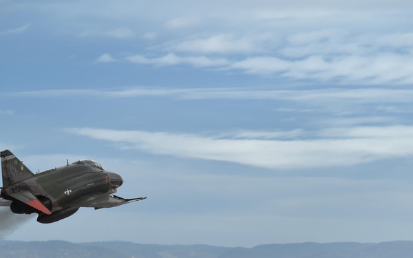 A military jet aircraft, identifiable as an F-4 Phantom II, is captured in a low-altitude, high-speed pass over what appears to be a military air base. The aircraft is in a banked turn with engine exhaust smoke visible, set against a desert landscape with mountains in the background. Ground infrastructure including hangars and antenna towers is visible below.