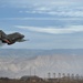 A military jet aircraft, identifiable as an F-4 Phantom II, is captured in a low-altitude, high-speed pass over what appears to be a military air base. The aircraft is in a banked turn with engine exhaust smoke visible, set against a desert landscape with mountains in the background. Ground infrastructure including hangars and antenna towers is visible below.