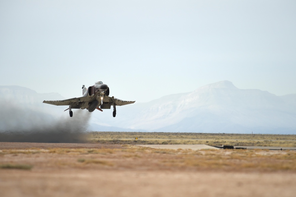 A military jet aircraft, consistent with an F-4 Phantom II, is captured at the moment of takeoff from a desert airstrip. The aircraft's landing gear is still extended and engine exhaust/heat distortion is visible behind it. The setting appears to be an arid, high-desert environment with mountains visible in the background.
