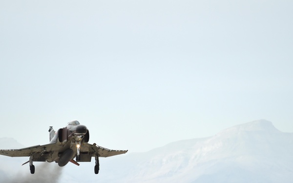 A military jet aircraft, consistent with an F-4 Phantom II, is captured at the moment of takeoff from a desert airstrip. The aircraft's landing gear is still extended and engine exhaust/heat distortion is visible behind it. The setting appears to be an arid, high-desert environment with mountains visible in the background.