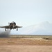 A military jet aircraft, consistent with an F-4 Phantom II, is captured at the moment of takeoff from a desert airstrip. The aircraft's landing gear is still extended and engine exhaust/heat distortion is visible behind it. The setting appears to be an arid, high-desert environment with mountains visible in the background.