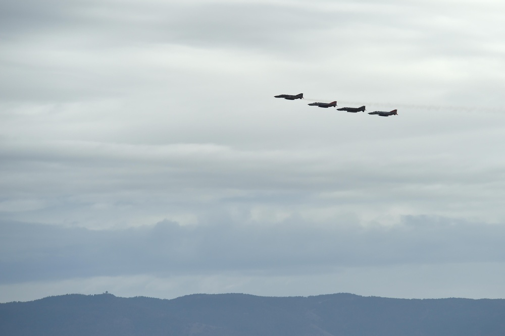 Four military jet aircraft are flying in a diagonal echelon formation against an overcast sky. The jets appear to have red and dark markings visible on their fuselages, suggesting they may be display or ceremonial aircraft. A low mountain or hill ridgeline is visible along the bottom of the frame beneath the formation.
