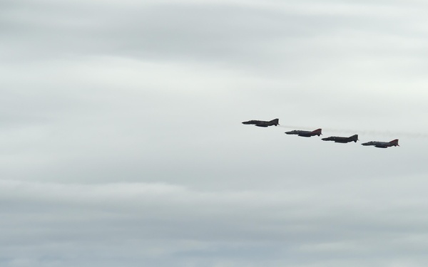 Four military jet aircraft are flying in a diagonal echelon formation against an overcast sky. The jets appear to have red and dark markings visible on their fuselages, suggesting they may be display or ceremonial aircraft. A low mountain or hill ridgeline is visible along the bottom of the frame beneath the formation.