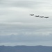 Four military jet aircraft are flying in a diagonal echelon formation against an overcast sky. The jets appear to have red and dark markings visible on their fuselages, suggesting they may be display or ceremonial aircraft. A low mountain or hill ridgeline is visible along the bottom of the frame beneath the formation.