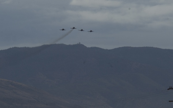 An air show scene featuring a formation of four military jets flying in close formation with a smoke trail against an overcast sky and mountainous backdrop. Two spectators are standing on top of a static display aircraft — one in military camouflage uniform, one in civilian clothing — watching and photographing the flyover. A crowd of additional spectators is visible in the foreground.