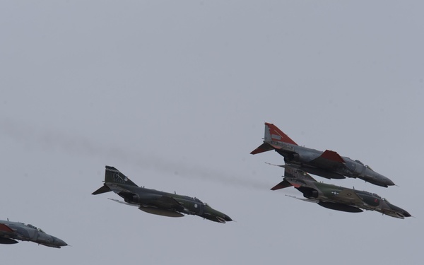 Four military jet aircraft are flying in a loose formation against an overcast gray sky. The aircraft appear to be F-4 Phantom II jets, with at least two displaying distinctive red and white markings on their tails and wingtips. One aircraft is producing a visible exhaust trail.