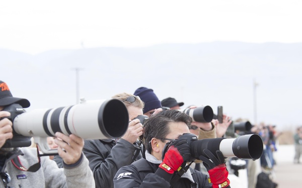 A group of photographers lined up on what appears to be an airfield tarmac, all pointing their cameras in the same direction. Several are using large white telephoto lenses, consistent with professional sports or aviation photography. The setting appears to be an outdoor media or press event in cool weather conditions.
