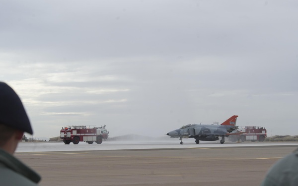 Two military personnel wearing berets observe a jet aircraft on a runway being sprayed with water by two red fire trucks positioned on either side. The aircraft appears to be an F-4 Phantom with an orange tail section, suggesting this may be a retirement or final flight ceremony known as a 'water salute.' The scene takes place on a military airfield under overcast skies.