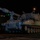 A self-propelled artillery piece, consistent with an M109 Paladin howitzer, is photographed at night on what appears to be a port or dock facility. The vehicle is painted in a desert tan/sand color scheme, suggesting recent or current deployment in an arid environment. A large vessel with green lighting is visible in the background, indicating a military logistics or sealift operation.