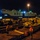 A nighttime military logistics operation at a port facility showing the loading or unloading of heavy military vehicles onto or from an ARC (American Roll-on Roll-off Carrier) vessel. Multiple armored military vehicles, including what appears to be an M1 Abrams tank and M113 armored personnel carriers, are staged on rail flatcars or dock platforms. Port workers in high-visibility orange safety gear are overseeing the operation.