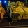 A nighttime photograph showing multiple tracked military armored fighting vehicles, likely M2 Bradley Infantry Fighting Vehicles, parked closely together on what appears to be a port or staging area. The vehicles are painted in desert tan/camouflage coloring and are illuminated by artificial lighting. Visible markings include unit identifiers and vehicle numbers such as 'D0035', 'D0036', 'WAOJAO', '410 588 BEB', and 'SGTCLINTON'.