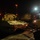 A military armored vehicle, appearing to be a self-propelled artillery piece or armored fighting vehicle, is photographed at night in what appears to be a port or logistics staging area. The vehicle is painted in a desert/tan color scheme and is positioned on cobblestone pavement near what appears to be a loading ramp or dock structure. The scene is illuminated by artificial lighting including orange sodium vapor lights and bright white floodlights.