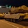 A nighttime scene showing multiple U.S. Army armored vehicles staged on a dock or pier alongside a large military cargo/transport ship. The foreground features what appears to be an M1 Abrams Armored Vehicle Launched Bridge (AVLB) variant with its bridge superstructure raised, alongside other tracked armored vehicles. The scene suggests a military loading or offloading operation at a port facility.