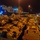 A nighttime port scene showing a large number of military armored vehicles, likely Bradley Fighting Vehicles, staged on a dock adjacent to a large roll-on/roll-off (RoRo) cargo vessel marked 'ARC'. The vehicles appear to be in desert tan/sand camouflage paint, suggesting preparation for deployment or return from an arid-region operation. The scene indicates a large-scale military logistics or sealift operation.
