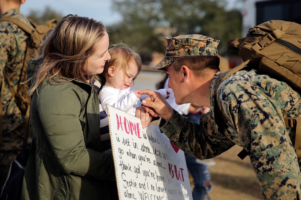 A U.S. Marine in woodland MARPAT camouflage uniform returns home and greets a young child being held by a woman, presumably his family. A handmade welcome home sign is visible, partially readable with the word 'HOME' and names including 'Sophie.' The scene captures an emotional military homecoming reunion.