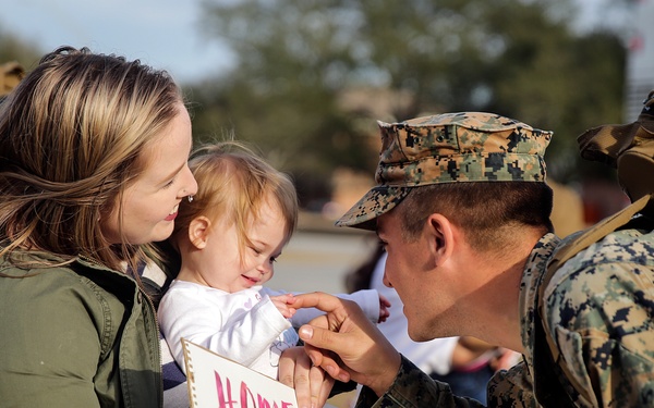 A U.S. Marine in woodland MARPAT camouflage uniform returns home and greets a young child being held by a woman, presumably his family. A handmade welcome home sign is visible, partially readable with the word 'HOME' and names including 'Sophie.' The scene captures an emotional military homecoming reunion.
