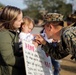 A U.S. Marine in woodland MARPAT camouflage uniform returns home and greets a young child being held by a woman, presumably his family. A handmade welcome home sign is visible, partially readable with the word 'HOME' and names including 'Sophie.' The scene captures an emotional military homecoming reunion.