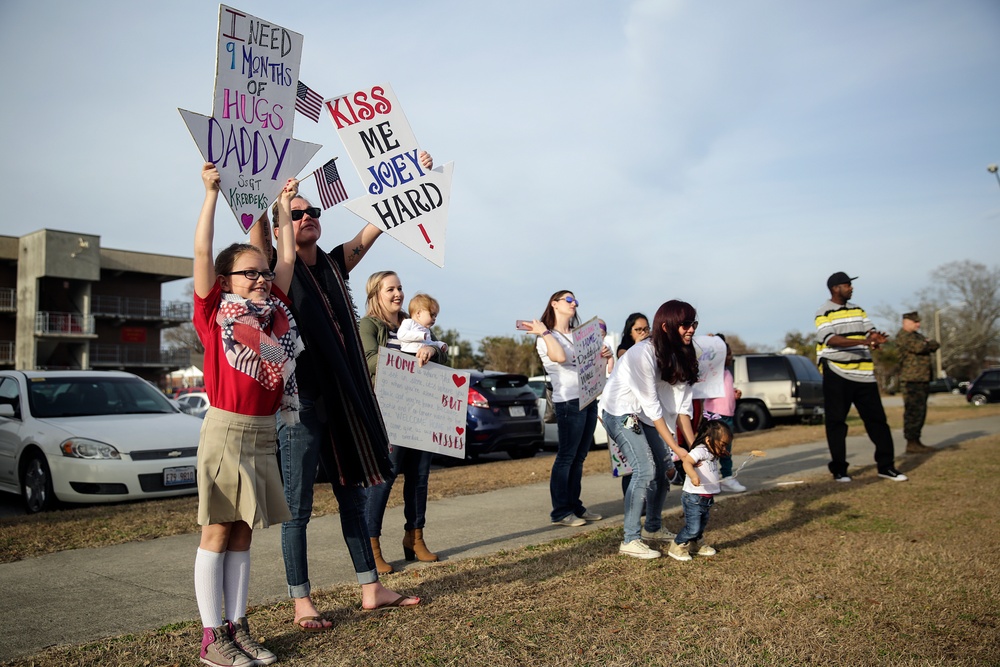 A group of family members, including children and women, are gathered outdoors holding handmade welcome-home signs, indicating a military homecoming event. The signs reference a deployment of approximately 9 months and express affection for returning service members. The setting appears to be a military base parking area, with at least one uniformed Marine visible in the background.