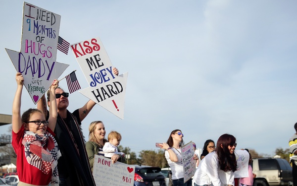 A group of family members, including children and women, are gathered outdoors holding handmade welcome-home signs, indicating a military homecoming event. The signs reference a deployment of approximately 9 months and express affection for returning service members. The setting appears to be a military base parking area, with at least one uniformed Marine visible in the background.