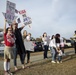 A group of family members, including children and women, are gathered outdoors holding handmade welcome-home signs, indicating a military homecoming event. The signs reference a deployment of approximately 9 months and express affection for returning service members. The setting appears to be a military base parking area, with at least one uniformed Marine visible in the background.