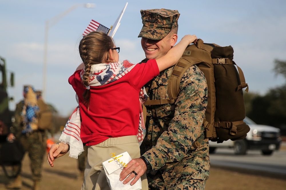 A U.S. Marine in woodland MARPAT camouflage uniform and cover is being embraced by a young girl wearing a red hoodie and an American flag-themed scarf. The Marine is smiling broadly and carrying a large tactical backpack, suggesting a homecoming or return from deployment. Small American flags are visible in the background along with other military personnel.
