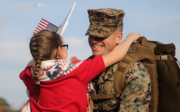 A U.S. Marine in woodland MARPAT camouflage uniform and cover is being embraced by a young girl wearing a red hoodie and an American flag-themed scarf. The Marine is smiling broadly and carrying a large tactical backpack, suggesting a homecoming or return from deployment. Small American flags are visible in the background along with other military personnel.