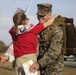 A U.S. Marine in woodland MARPAT camouflage uniform and cover is being embraced by a young girl wearing a red hoodie and an American flag-themed scarf. The Marine is smiling broadly and carrying a large tactical backpack, suggesting a homecoming or return from deployment. Small American flags are visible in the background along with other military personnel.