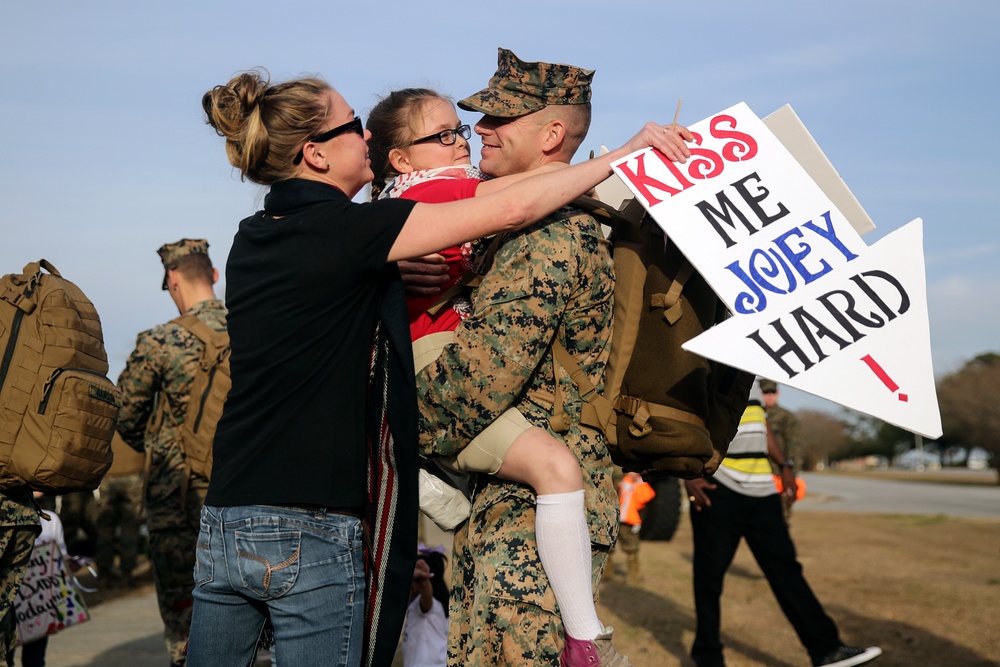 A U.S. Marine in camouflage uniform is being greeted by a woman and a young girl, likely his family, during what appears to be a military homecoming event. The marine is holding the child while embracing the woman, and a handmade arrow-shaped sign reads 'KISS ME JOEY HARD!' Other Marines and civilians are visible in the background.