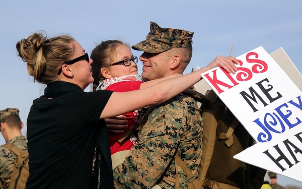 A U.S. Marine in camouflage uniform is being greeted by a woman and a young girl, likely his family, during what appears to be a military homecoming event. The marine is holding the child while embracing the woman, and a handmade arrow-shaped sign reads 'KISS ME JOEY HARD!' Other Marines and civilians are visible in the background.
