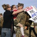 A U.S. Marine in camouflage uniform is being greeted by a woman and a young girl, likely his family, during what appears to be a military homecoming event. The marine is holding the child while embracing the woman, and a handmade arrow-shaped sign reads 'KISS ME JOEY HARD!' Other Marines and civilians are visible in the background.