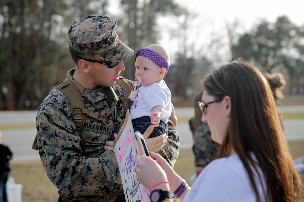 A U.S. Marine in digital camouflage uniform, identified by the name tape 'HOLLOWAY,' holds a young infant wearing a purple headband and pacifier at what appears to be a military homecoming event. A woman, likely the spouse, stands nearby holding a 'Welcome Home Daddy' sign and a phone, presumably taking photos. The scene captures an emotional family reunion moment outdoors on a military installation.