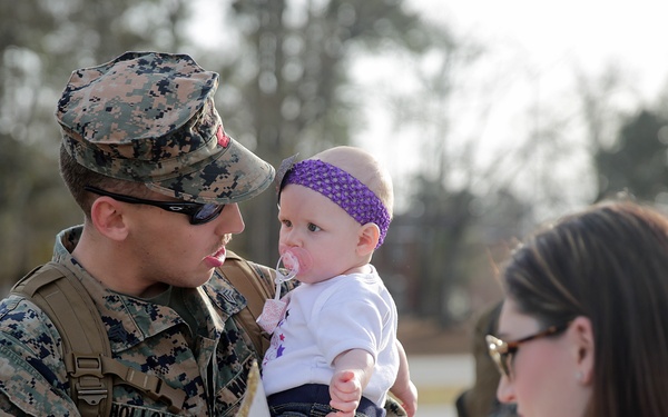 A U.S. Marine in digital camouflage uniform, identified by the name tape 'HOLLOWAY,' holds a young infant wearing a purple headband and pacifier at what appears to be a military homecoming event. A woman, likely the spouse, stands nearby holding a 'Welcome Home Daddy' sign and a phone, presumably taking photos. The scene captures an emotional family reunion moment outdoors on a military installation.