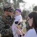 A U.S. Marine in digital camouflage uniform, identified by the name tape 'HOLLOWAY,' holds a young infant wearing a purple headband and pacifier at what appears to be a military homecoming event. A woman, likely the spouse, stands nearby holding a 'Welcome Home Daddy' sign and a phone, presumably taking photos. The scene captures an emotional family reunion moment outdoors on a military installation.