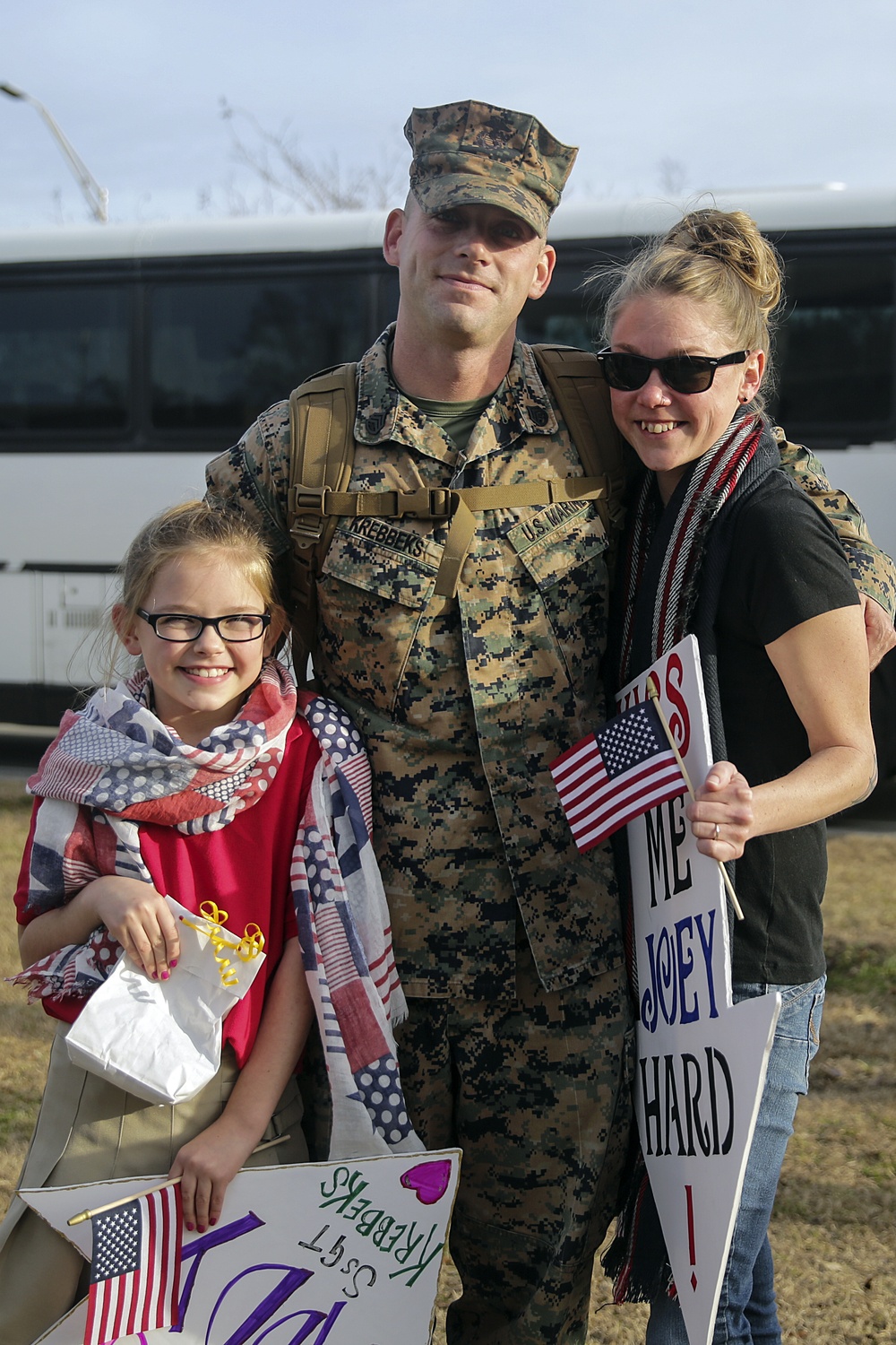 A U.S. Marine in MARPAT camouflage uniform, identified by name tape as 'Krebbeks', poses with a woman and a young girl in what appears to be a homecoming reunion. The group is smiling and holding American flags and welcome signs. The setting appears to be an outdoor military installation with a white bus visible in the background.