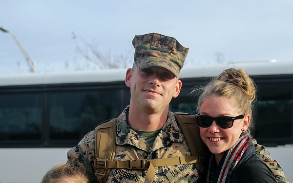 A U.S. Marine in MARPAT camouflage uniform, identified by name tape as 'Krebbeks', poses with a woman and a young girl in what appears to be a homecoming reunion. The group is smiling and holding American flags and welcome signs. The setting appears to be an outdoor military installation with a white bus visible in the background.