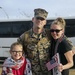 A U.S. Marine in MARPAT camouflage uniform, identified by name tape as 'Krebbeks', poses with a woman and a young girl in what appears to be a homecoming reunion. The group is smiling and holding American flags and welcome signs. The setting appears to be an outdoor military installation with a white bus visible in the background.