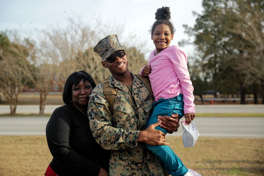 A U.S. Marine in full camouflage uniform, identified by the name tape 'PERRY,' poses with two family members outdoors on what appears to be a military base. He is holding a young girl in a pink top and teal pants while a woman in a black top stands beside him. All three are smiling broadly, suggesting a joyful reunion or family visit.