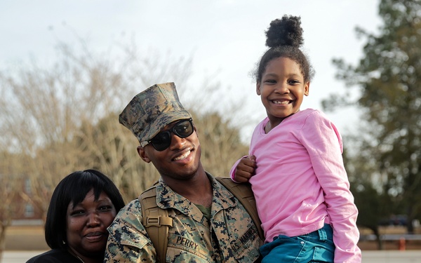 A U.S. Marine in full camouflage uniform, identified by the name tape 'PERRY,' poses with two family members outdoors on what appears to be a military base. He is holding a young girl in a pink top and teal pants while a woman in a black top stands beside him. All three are smiling broadly, suggesting a joyful reunion or family visit.