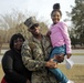A U.S. Marine in full camouflage uniform, identified by the name tape 'PERRY,' poses with two family members outdoors on what appears to be a military base. He is holding a young girl in a pink top and teal pants while a woman in a black top stands beside him. All three are smiling broadly, suggesting a joyful reunion or family visit.