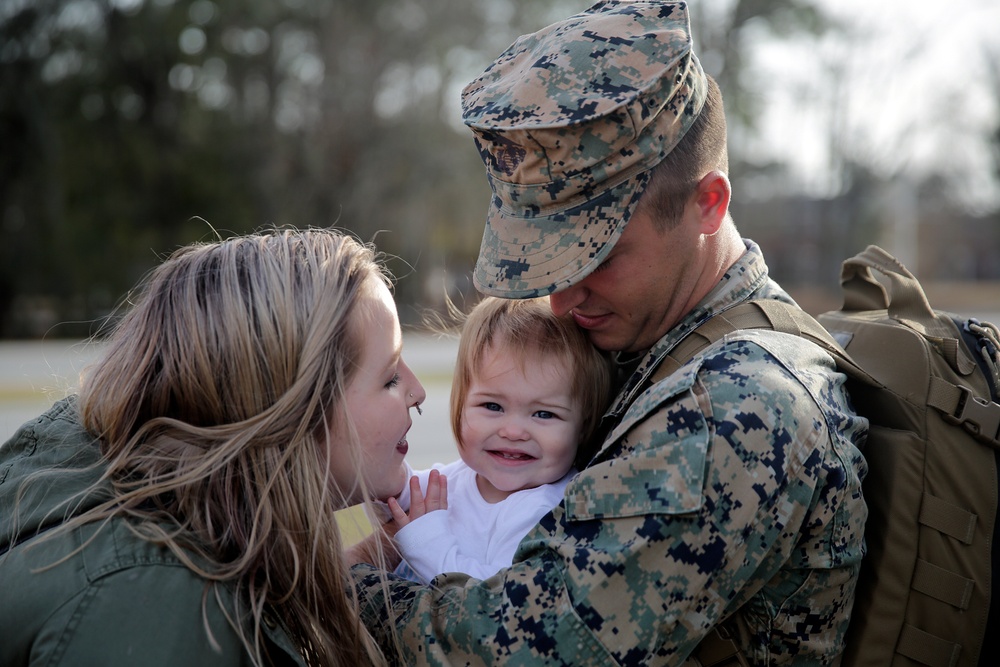 A U.S. Marine in full digital camouflage uniform and cover (cap) embraces a young toddler while a woman, likely his spouse or partner, leans in closely. The toddler is smiling broadly, suggesting a joyful reunion scene, possibly a homecoming from deployment. The emotional moment is captured outdoors with a blurred natural background.